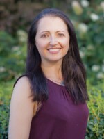 Headshot of a white middle aged woman with dark mid length hair and a burgundy sleeveless top. In the background are lush greenery and old university buildings.