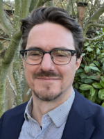 Closeup and headshot of a white middle-aged man in a blue blazer and wearing black glasses with brown hair. In the background, green trees and foliage are visible.