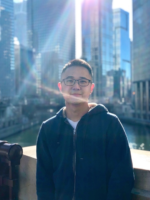 A younger man in a hoody standing in front of an urban landscape with the sun reflecting off the lens.